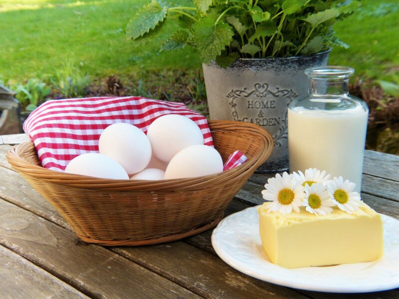 Fresh farm eggs in a wicker basket lined with a red and white checkered cloth, accompanied by a bottle of milk and a block of butter adorned with daisies, set on a wooden table with a green garden backdrop.